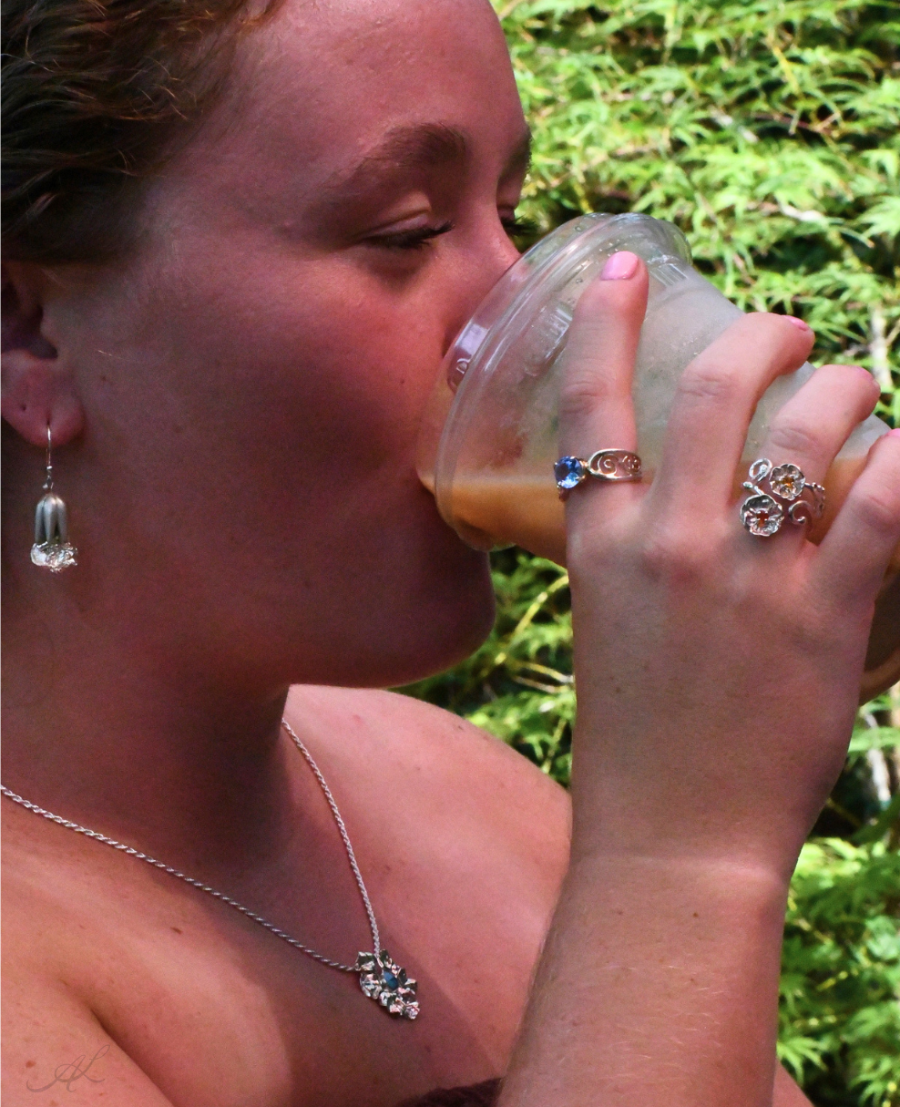 A close-up lifestyle shot of a woman drinking from a clear plastic cup outdoors, with green foliage in the background. She is wearing a silver necklace, earring, and rings.