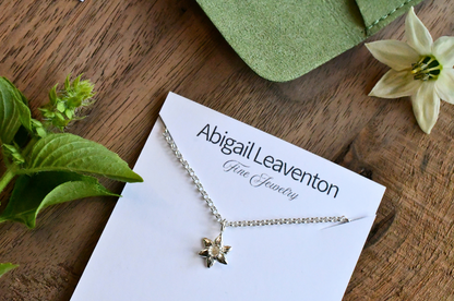 A delicate silver necklace with a small flower-shaped pendant displayed on a white card printed with the brand name 'Abigail Leaventon Fine Jewelry'. The card is styled on a rustic wooden surface with green leaves, a green pouch, and a white flower.