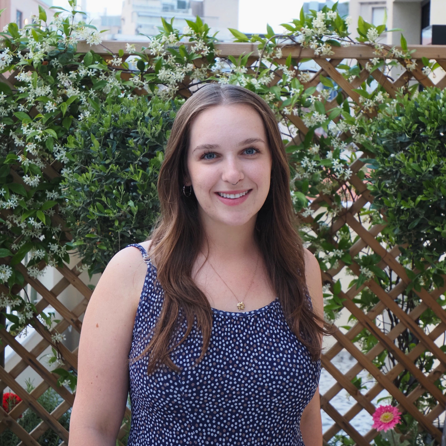 A smiling young woman with long brown hair stands outdoors in front of a wooden lattice fence covered in green foliage and small white flowers. She is wearing a sleeveless navy top with a small white polka dot pattern.