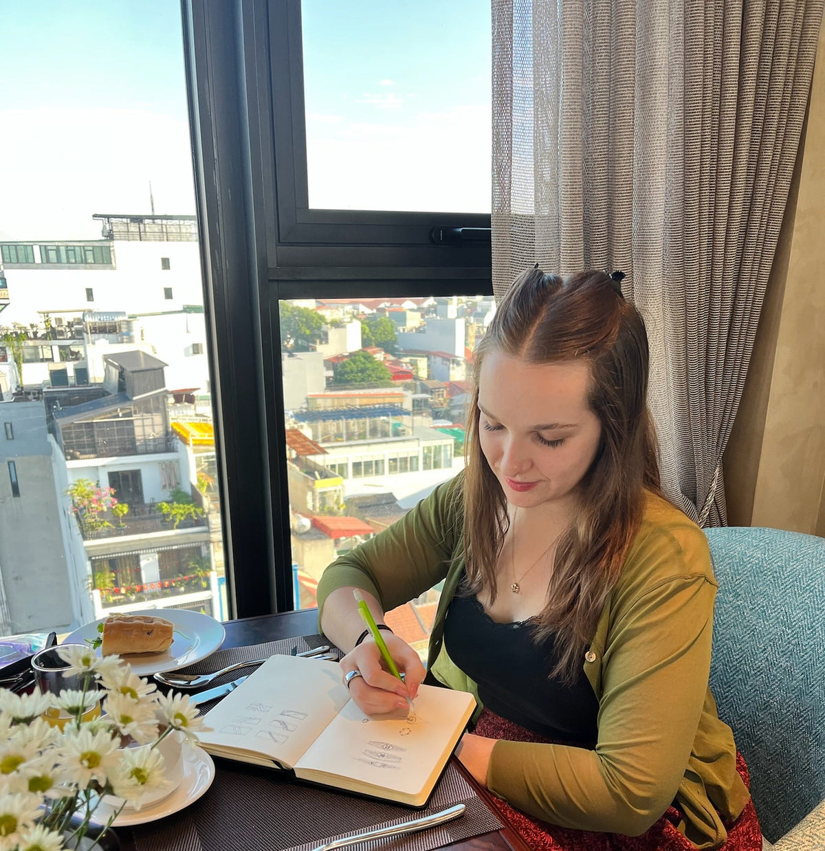 A woman sits at a wooden table by a large window, writing in an open notebook with a green pen. She is wearing a green cardigan over a black top and a red patterned skirt.
