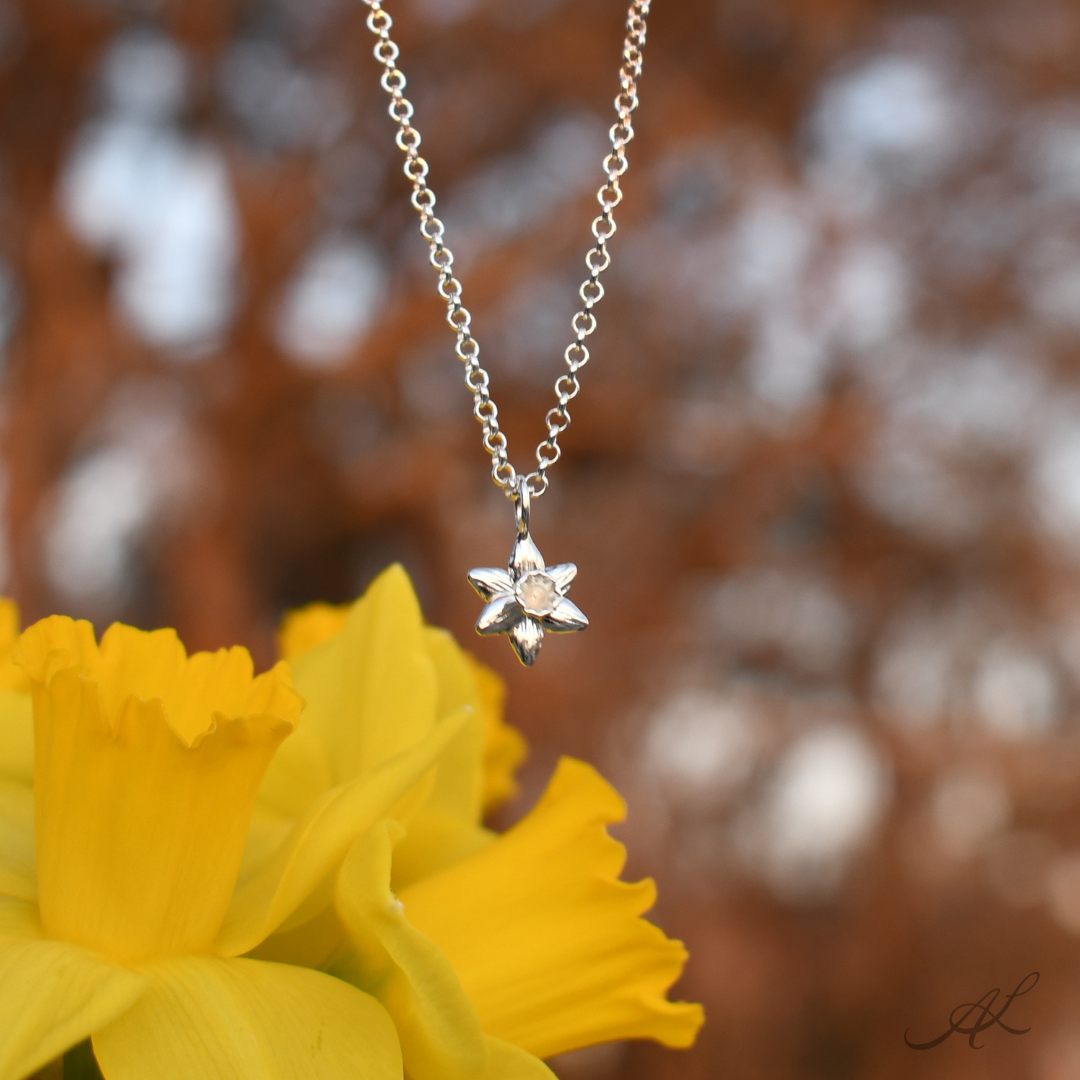 A silver six-pointed star pendant necklace on a delicate chain, displayed in front of a bright yellow daffodil flower with a blurred brown and white bokeh background.